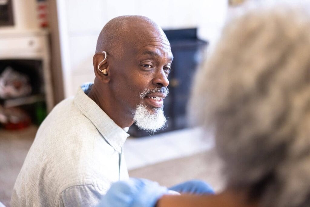 An older adult man sits, talking with a woman who is mostly out of frame. He is wearing a hearing aid.