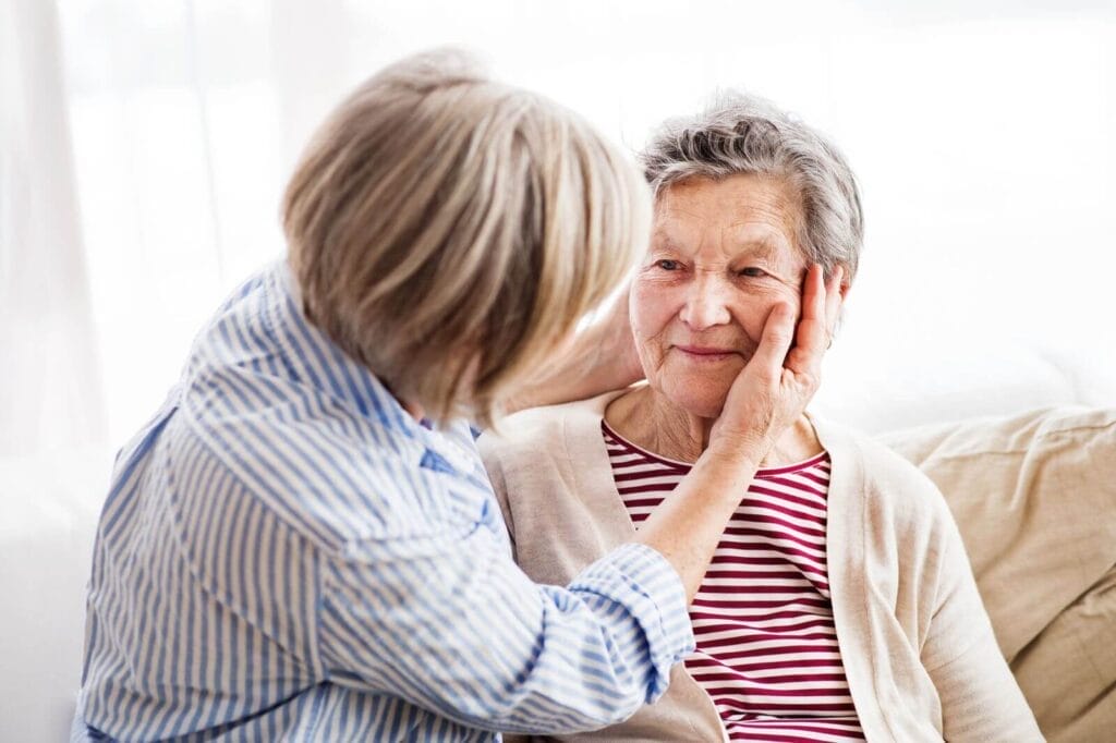 A woman with her back to the camera places her hands on the face of an older adult woman seated next to her.