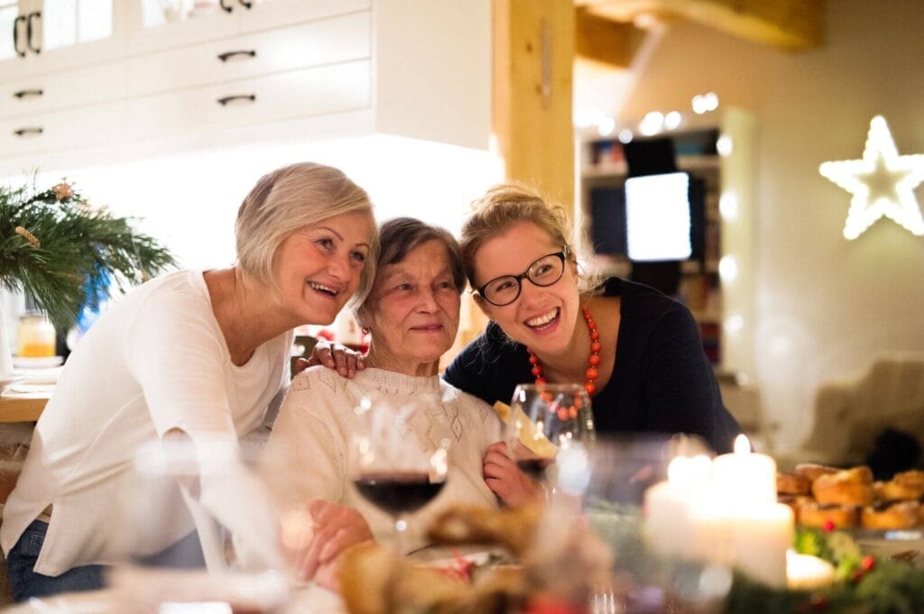 An older adult woman sits at a chair at a table full of food and drink. Two younger women are on either side of her. They appear to be smiling at a camera that is not pictured.