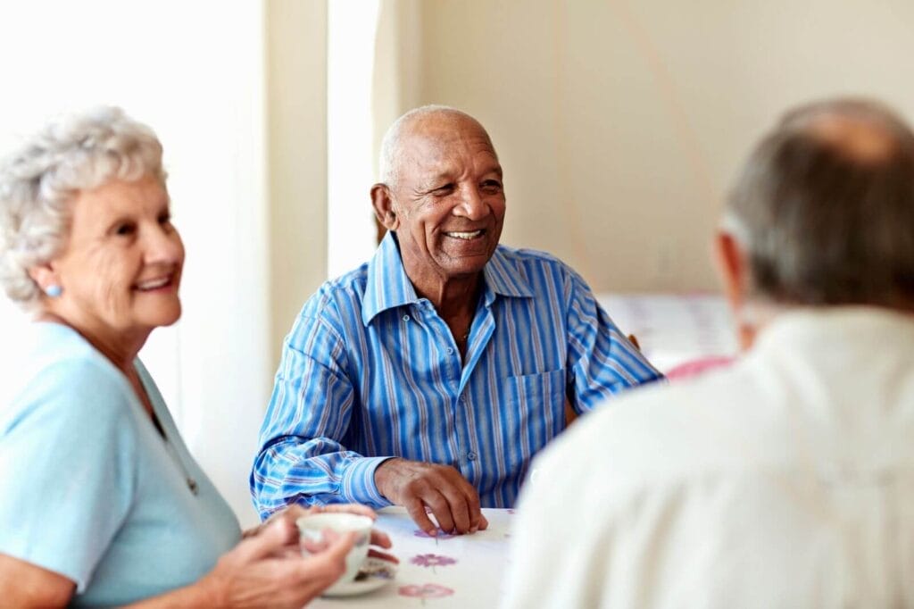 Two older adult men and one older adult woman sit at a table. The woman has a teacup. They smile.