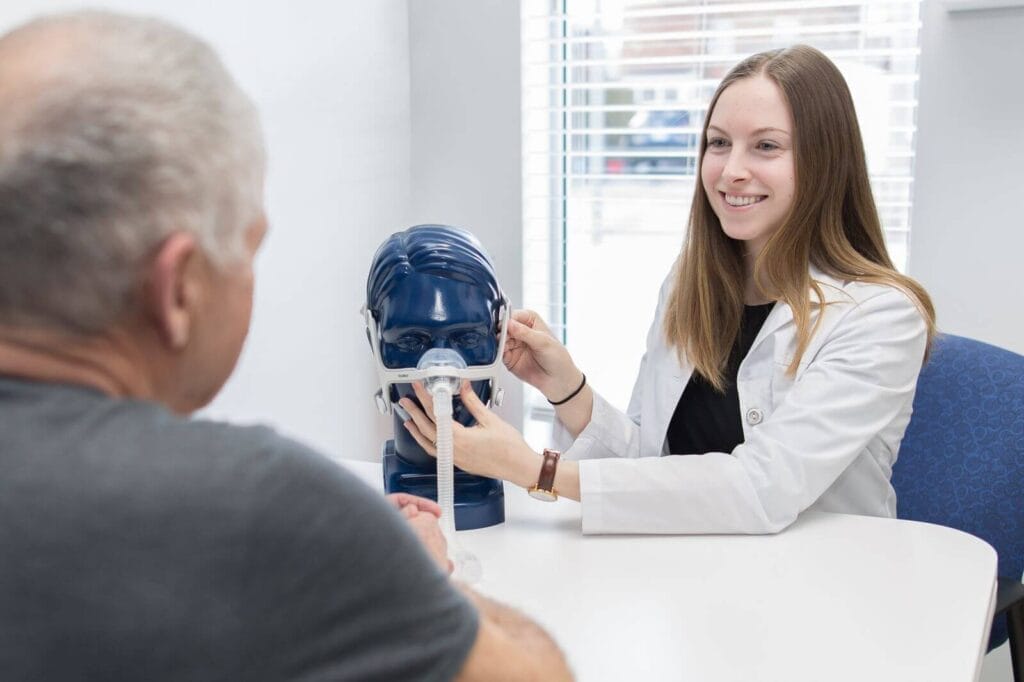 A woman wearing a lab coat shows an older adult man how to use a CPAP machine. She has placed the face mask on a mannequin head to demonstrate.