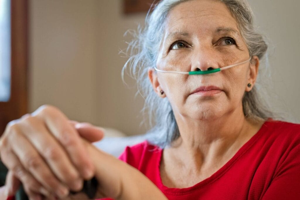 An older adult woman sits with a cannula for oxygen in her nose.