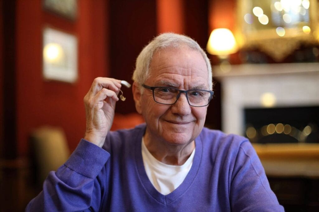 An older adult man holds a hearing aid in his hand, about to place it in his ear.