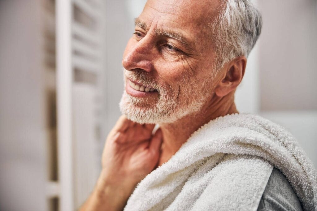 An older adult man with a bath towel over his shoulder smiles at something off camera.