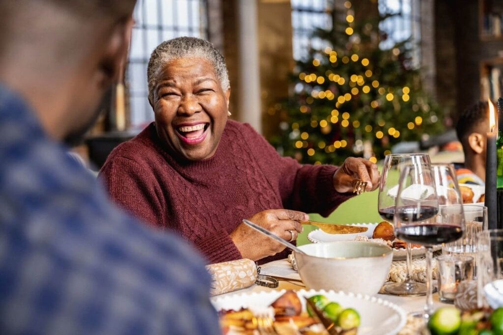 An older adult woman is sitting at the table for a holiday feast. She is laughing with the man sitting next to her. There is a Christmas tree in the background.