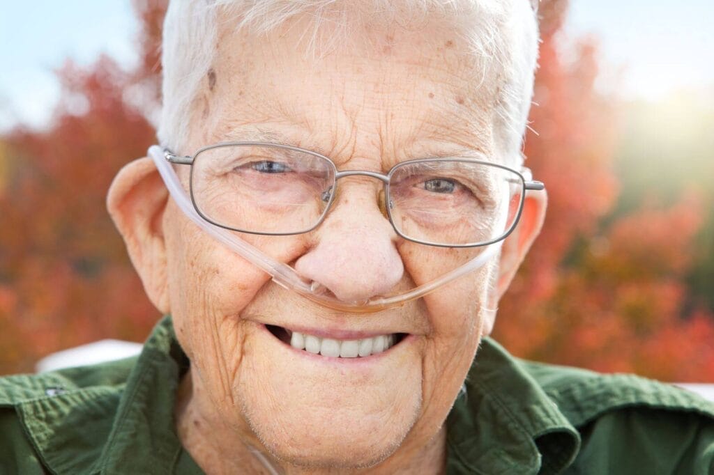 An older adult man with nasal prongs for oxygen smiles at the camera.