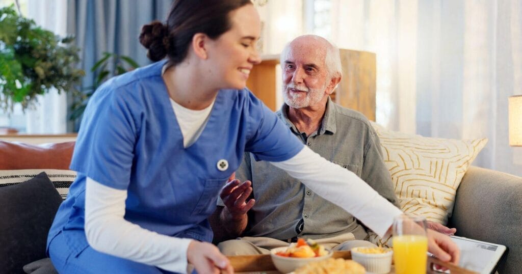 A female caregiver wearing scrubs places a tray of food in front of an older adult man seated on a couch.