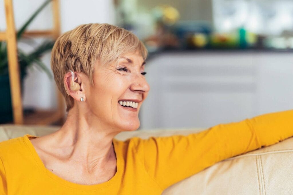 An older adult woman wearing a hearing aid is sitting on a couch, smiling at someone off camera.