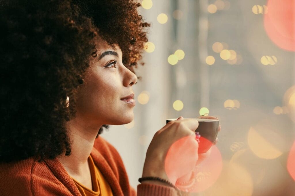 A woman is shown in profile drinking a cup of coffee. There are twinkly lights in the background.