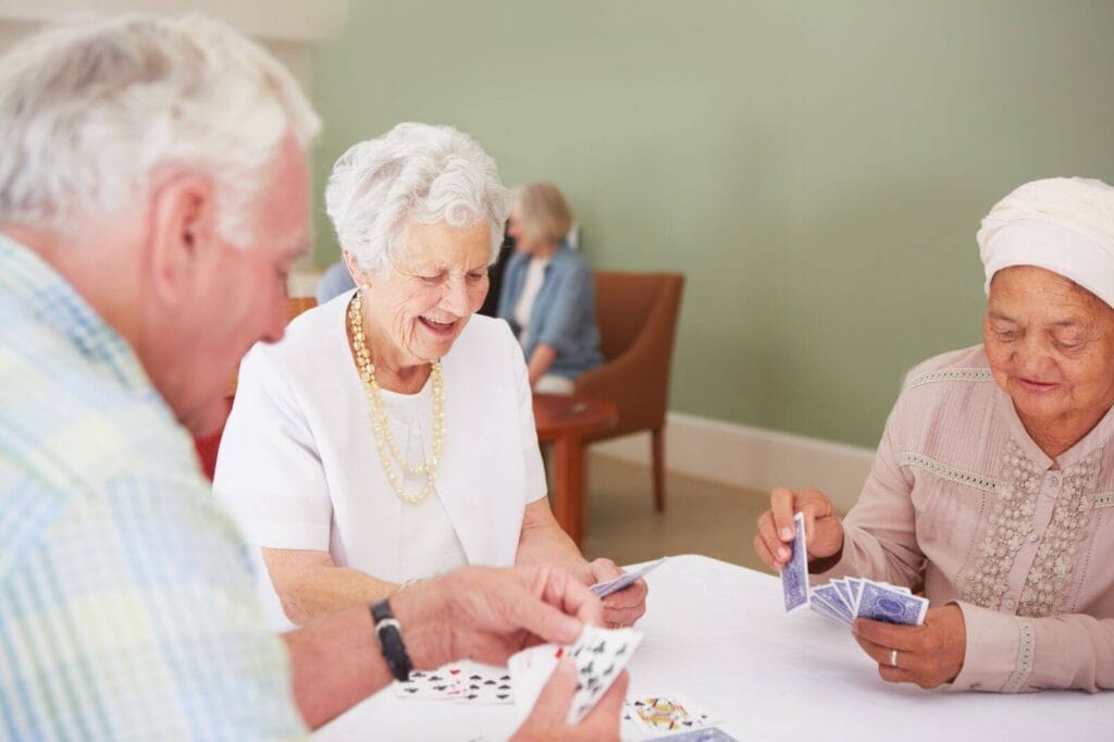 Two older adult women and one older adult man sit at a table playing cards. There is another older adult woman sitting on a chair in the background.