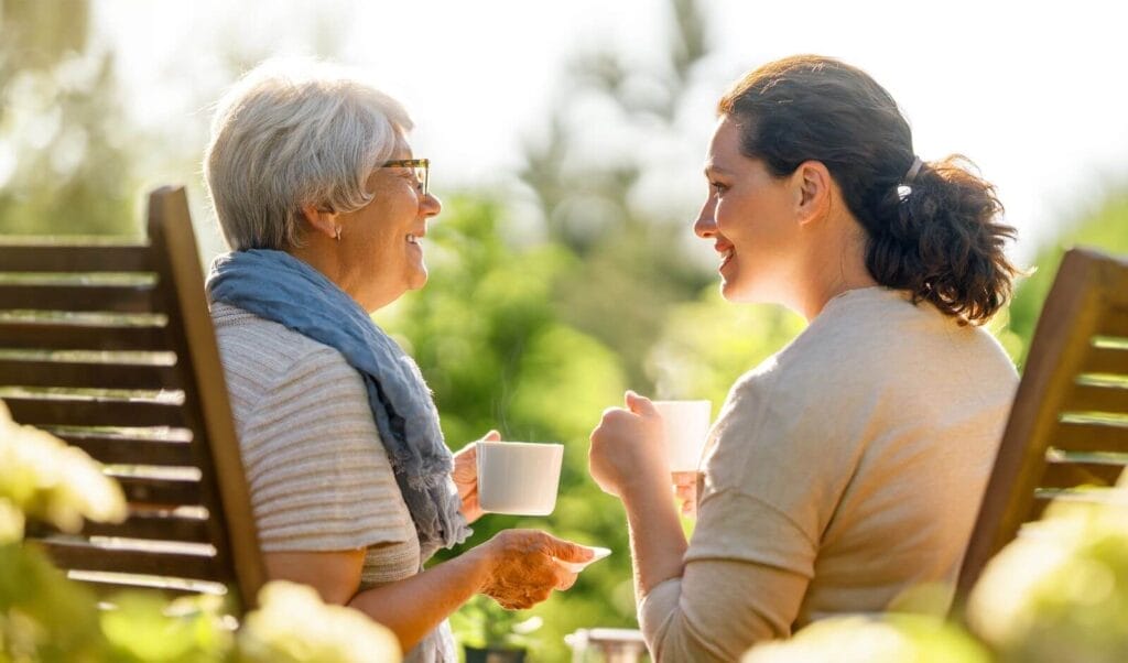 Shown from behind, a woman sits with outside with an older adult woman. They are in wooden chairs, holding coffee mugs, and smiling at each other.