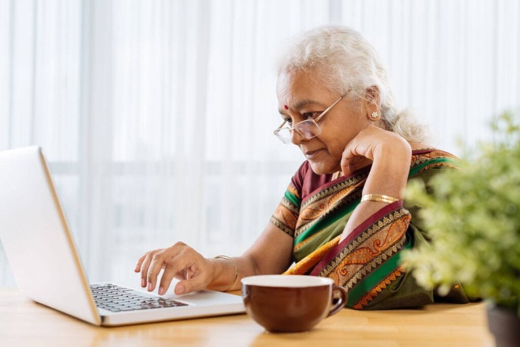 An older adult woman sits at a table with a laptop and a cup of coffee.