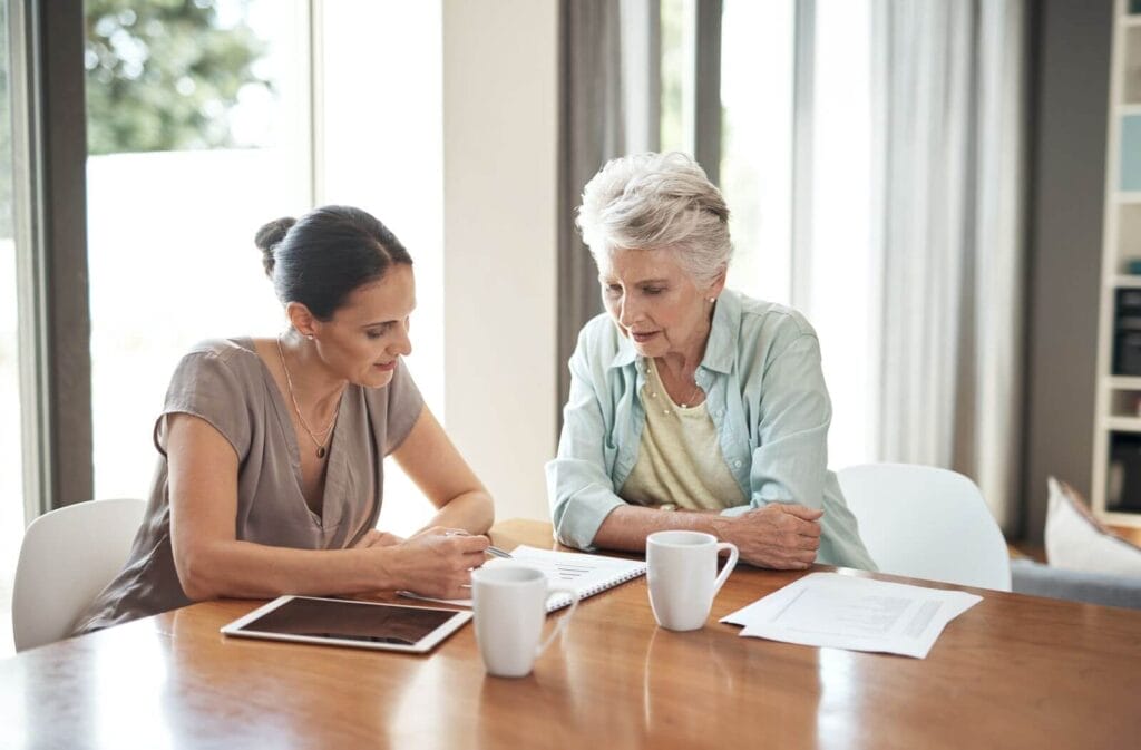 A woman and her older adult mother sit at a table looking over some paperwork.