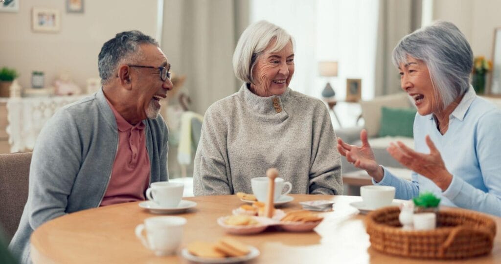 Two older adult women and one older adult man sit at a table with tea and cookies in front of them. They are talking and smiling.