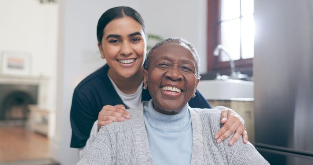 A female caregiver stands behind an older adult woman with her hands on her shoulders. They smile at the camera.