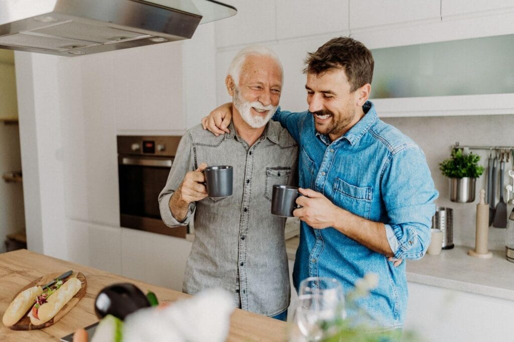 A man has his arm around the shoulder of his older adult father. They are holding coffee mugs and smiling.