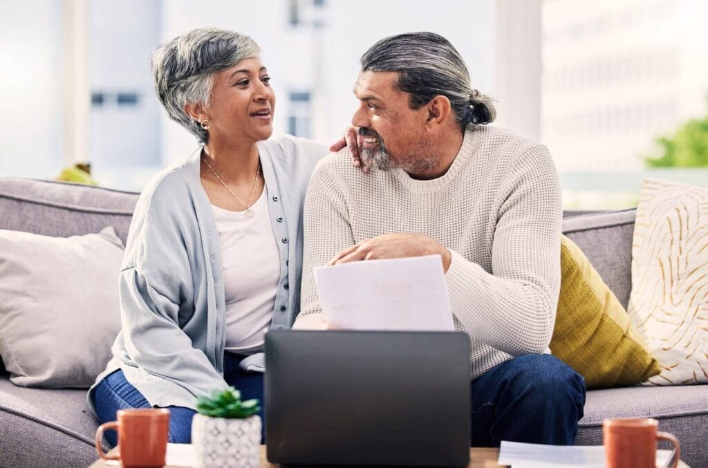 A man and woman sit on a couch, looking over paperwork. There is a laptop on the coffee table in front of them.