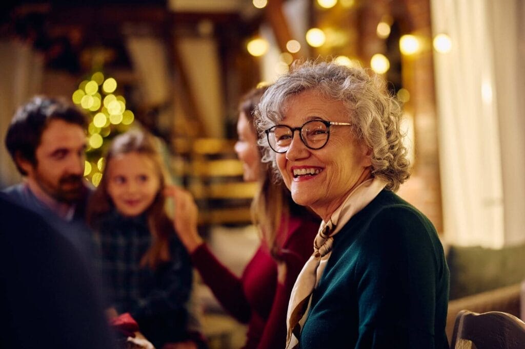 A smiling older adult woman sits with a man, woman, and their young daughter. There is a festive atmosphere.