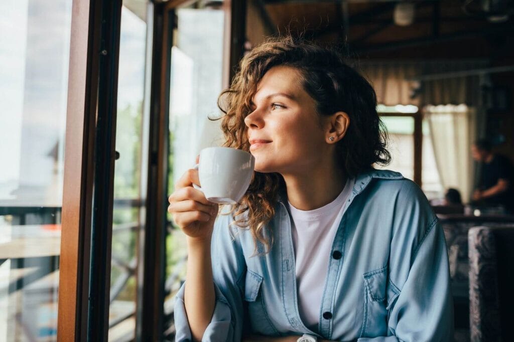 A woman sits at a table holding a coffee cup. She looks out a window and smiles.