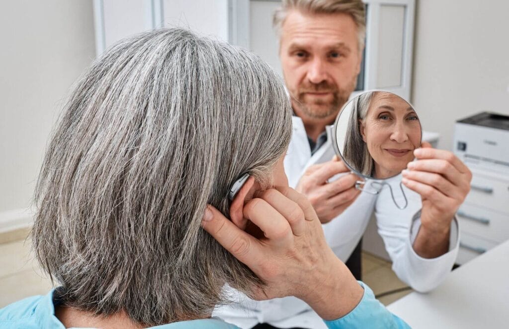 An audiologist holds a mirror up to an older adult woman who is trying on new hearing aids.