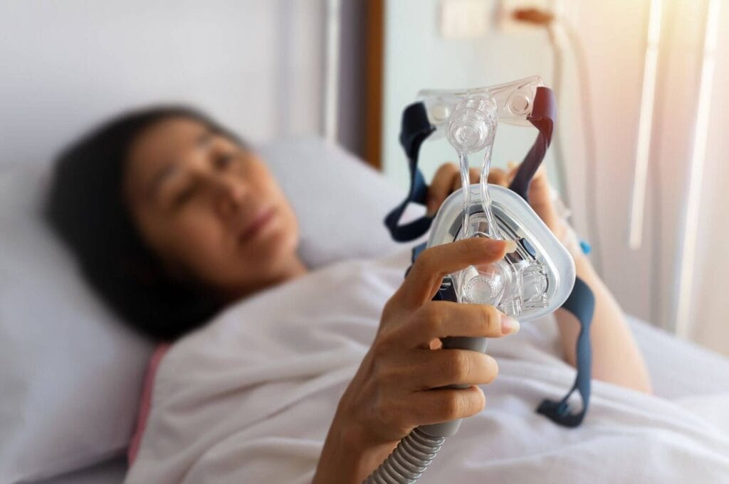 An older adult woman lies in bed holding a mask for a CPAP machine for sleep apnea.