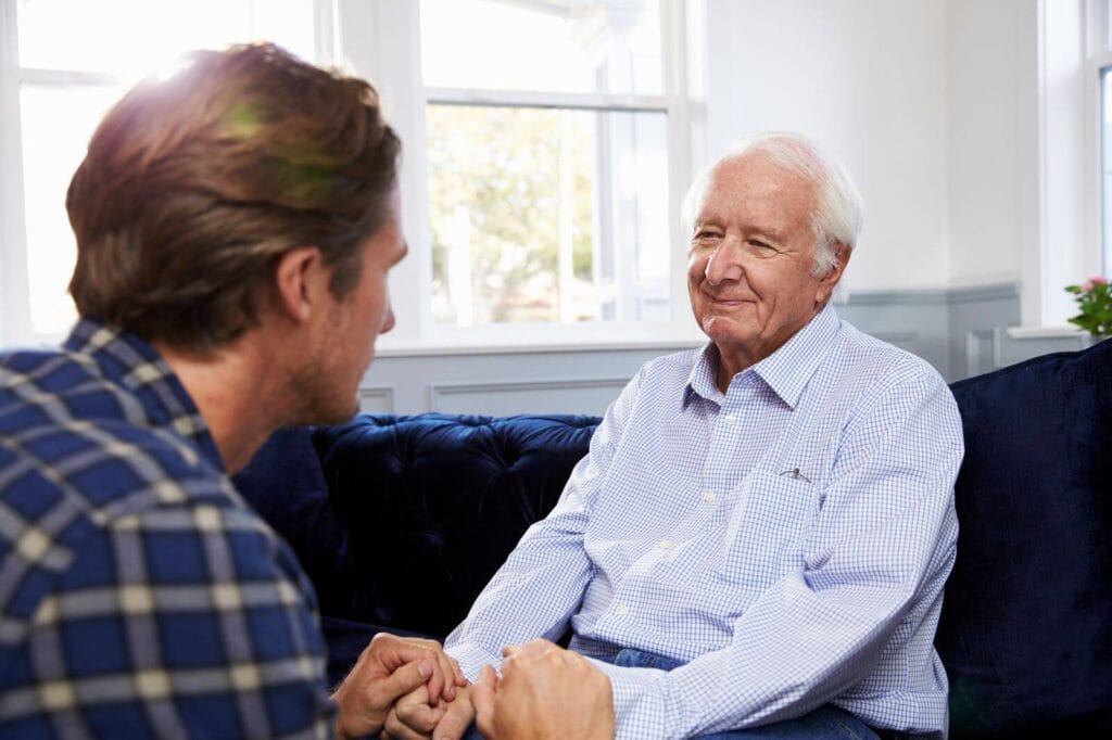 A man sits with his older adult father, holding his hands.