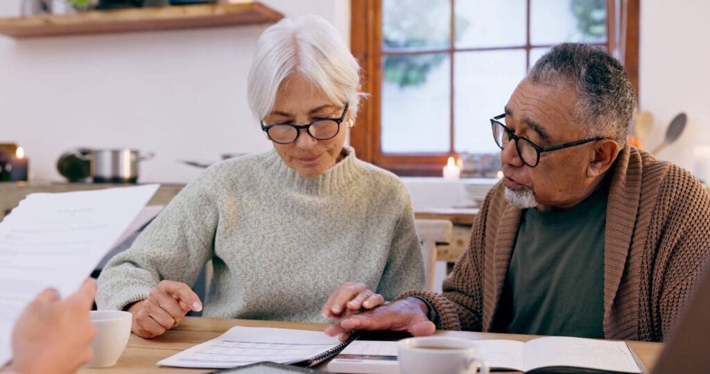 An older adult man and woman sit at a table looking over paperwork. There is a person seated across from them whose hand is shown holding more paperwork.