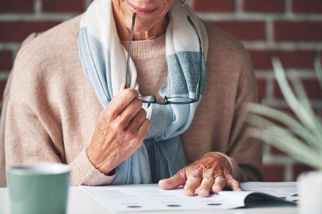 An older adult woman is holding reading glasses to her mouth. She is looking over some paperwork.