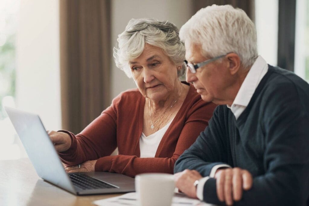 An older adult man and woman sit at a table looking at a laptop. There is a coffee mug and some paperwork on the table as well.