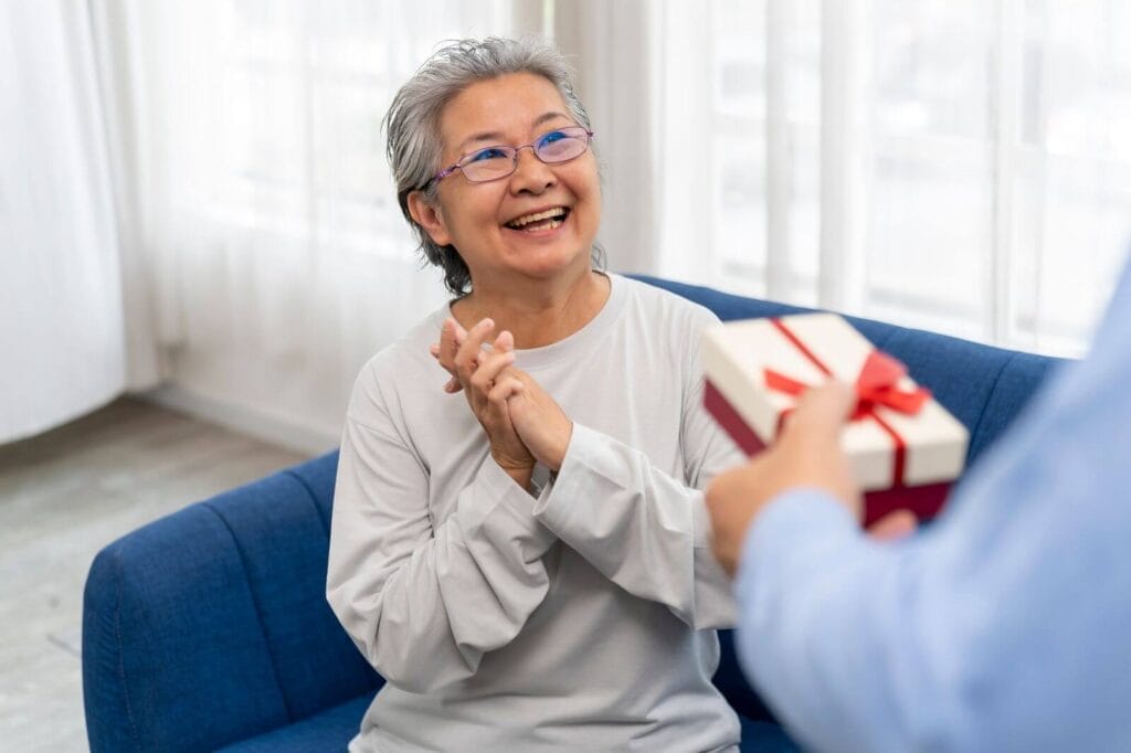 A happy older adult woman sits on a couch as someone off-camera hands her a gift with a bow on it.