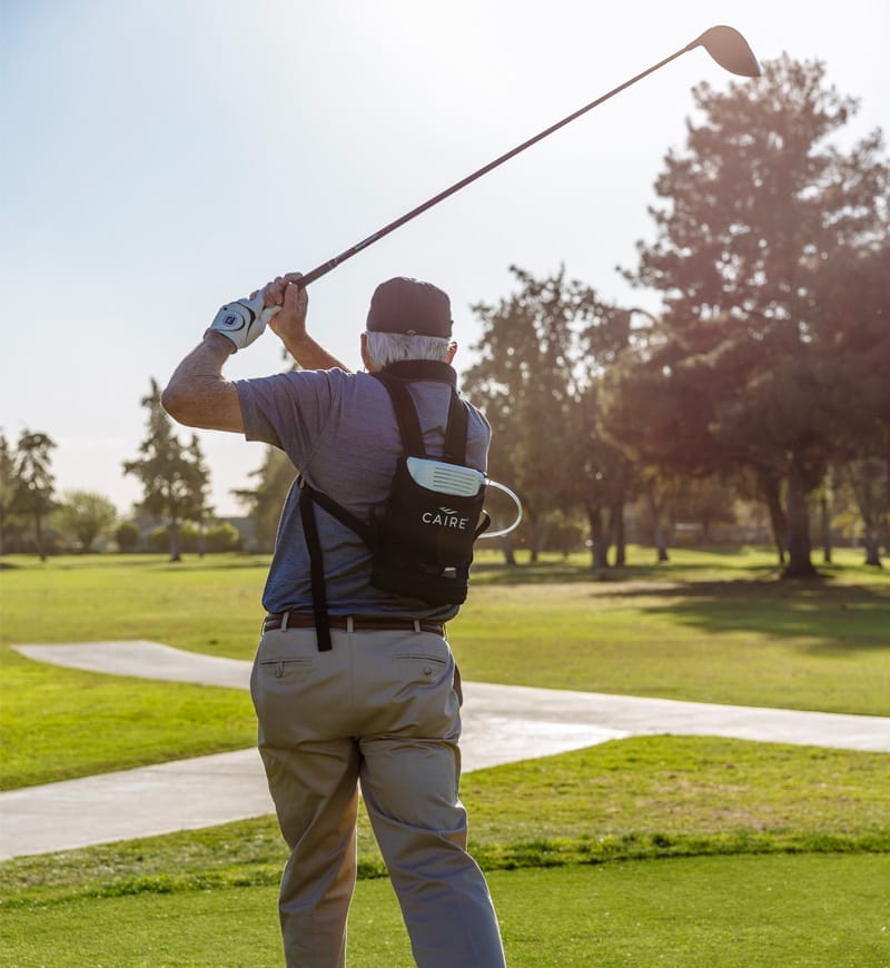 An image of a man who just swung a golf club watching the ball sail through the sky. He wears a portable oxygen concentrator on his back.