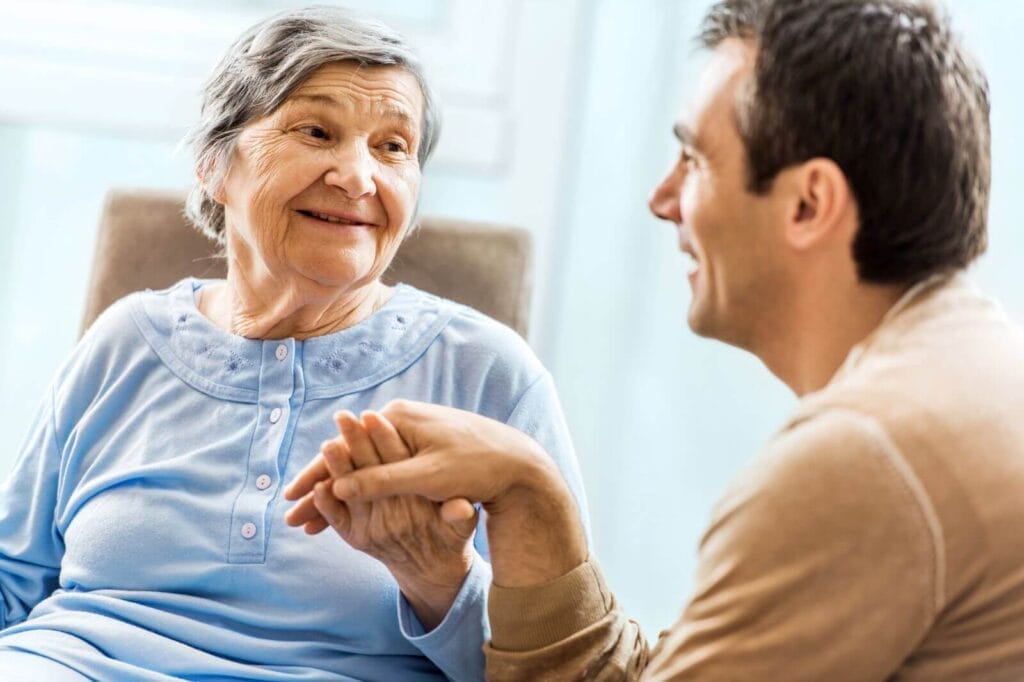 A man sits with his older adult mother and holds her hand. She smiles at him.