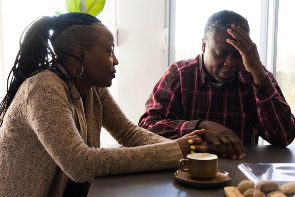 A woman sits at a table with her older adult father. They are discussing something. There is a coffee mug in front of her.