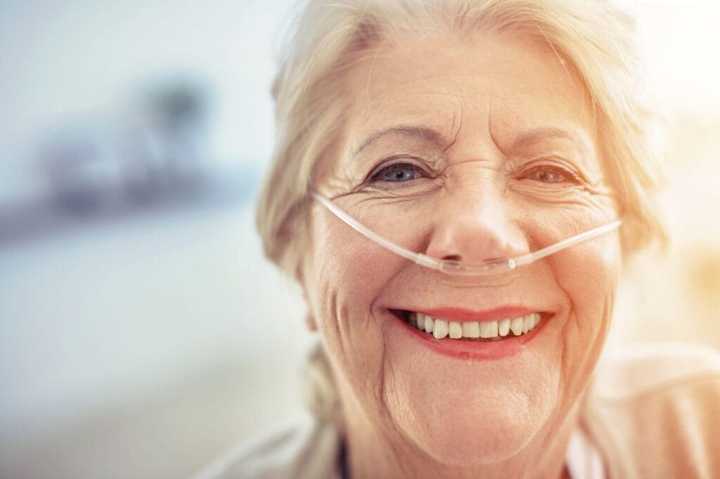 An older adult woman with a nasal cannula smiles at the camera.