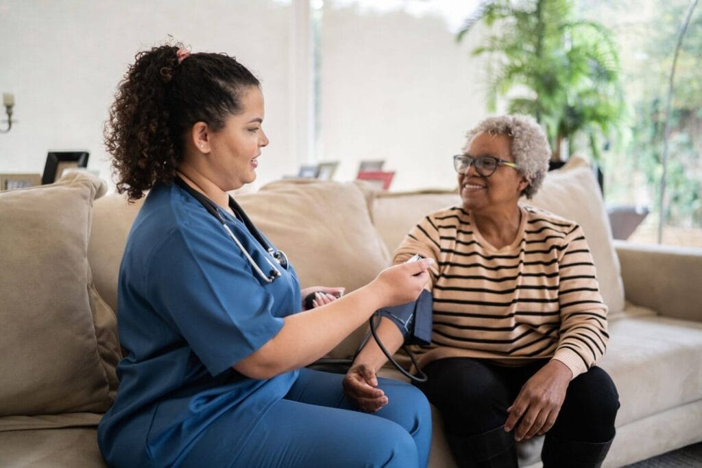 A female health care worker takes the blood pressure of an older adult woman as they sit on a couch in her home.