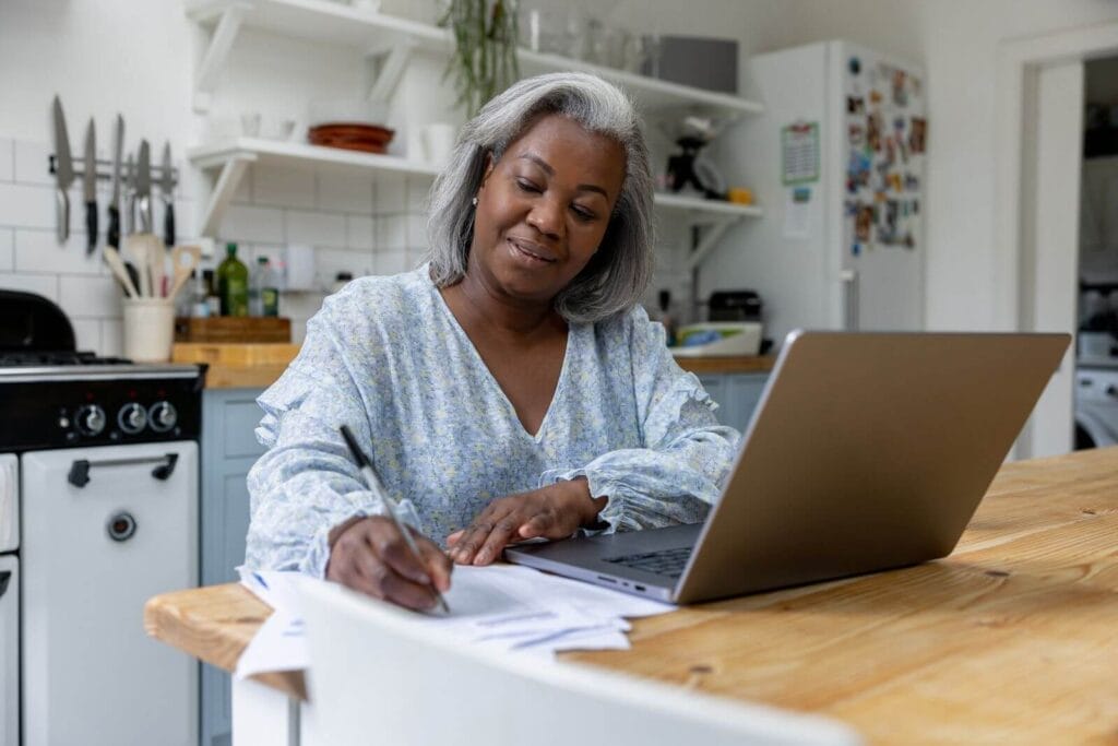 A woman sits at her kitchen table with a laptop in front of her. She is writing on some paperwork.