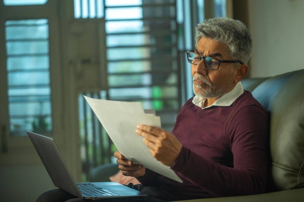 An older adult man sits on a couch with a laptop on his lap. He is looking at some paperwork.