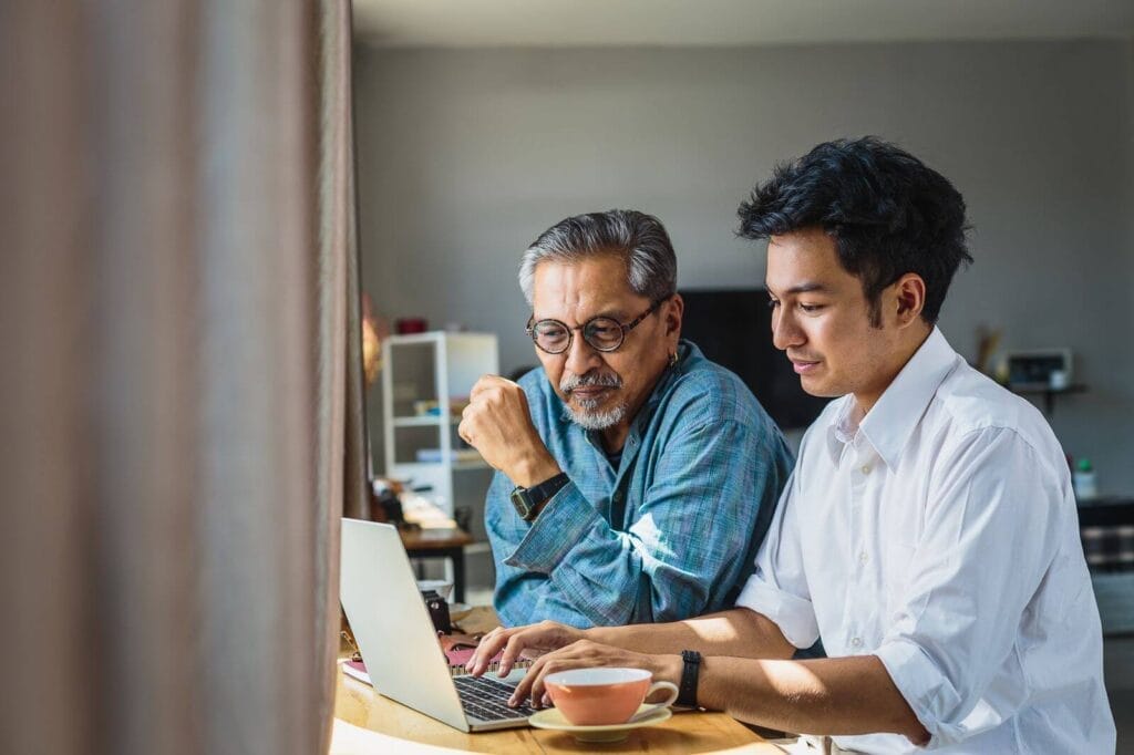 An older adult man and his son sit at a table looking at a laptop.