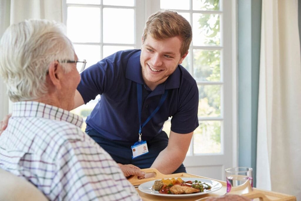 A male caregiver places a tray with a place of food and a glass of water in front of an older adult man.