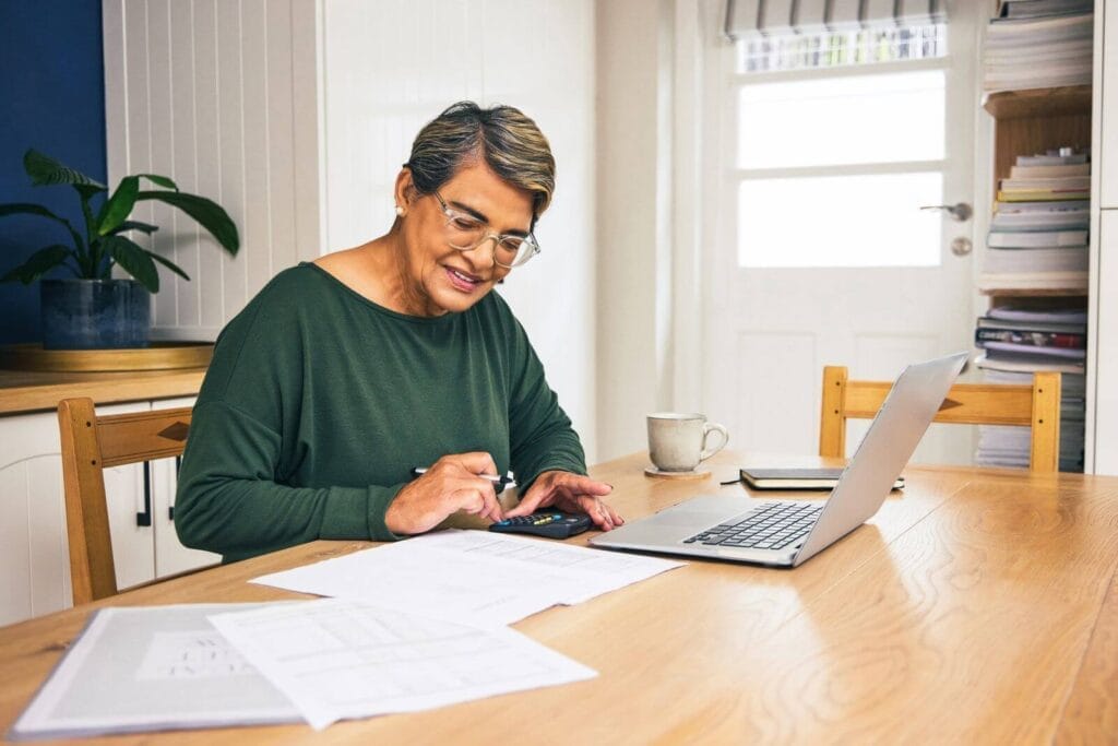 An older adult woman sits at at table with paperwork, a calculator, and a laptop in front of her.