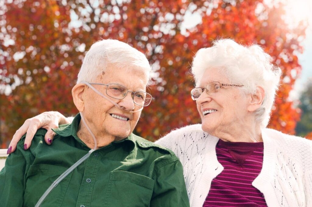 An older adult man and woman sit outside. The man is wearing a nasal cannula for oxygen. The woman has her arm around his shoulder.