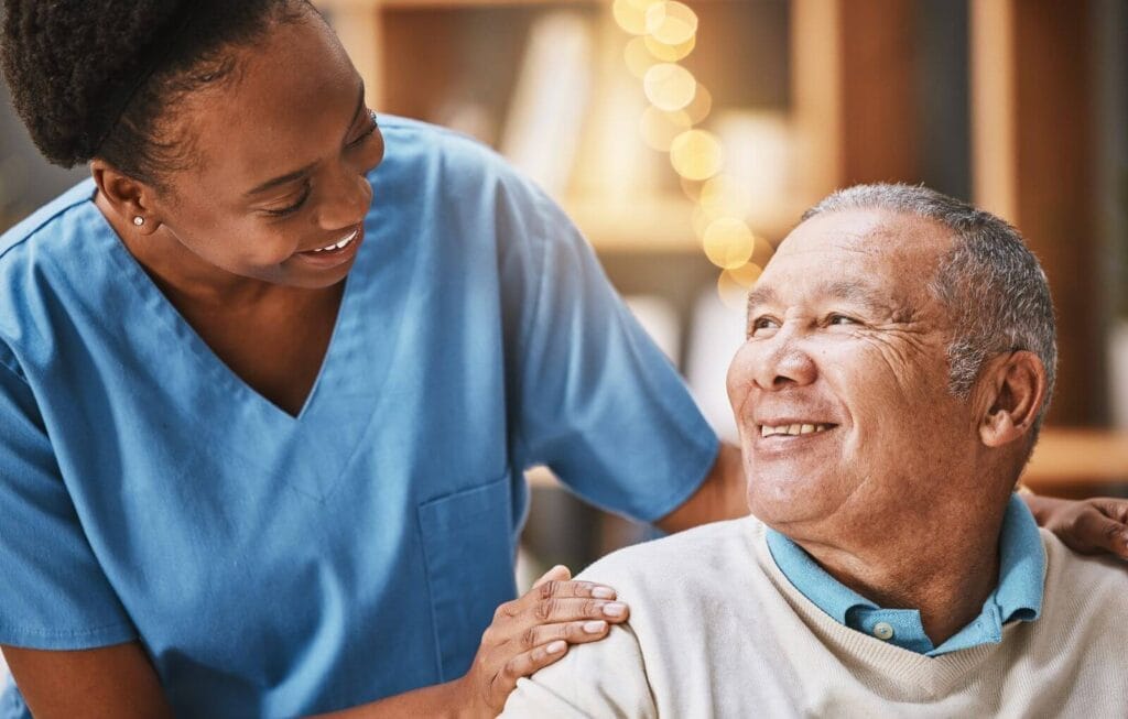 An older adult man is seated and smiling at a female caregiver, who stands behind him with her hands on his shoulders.