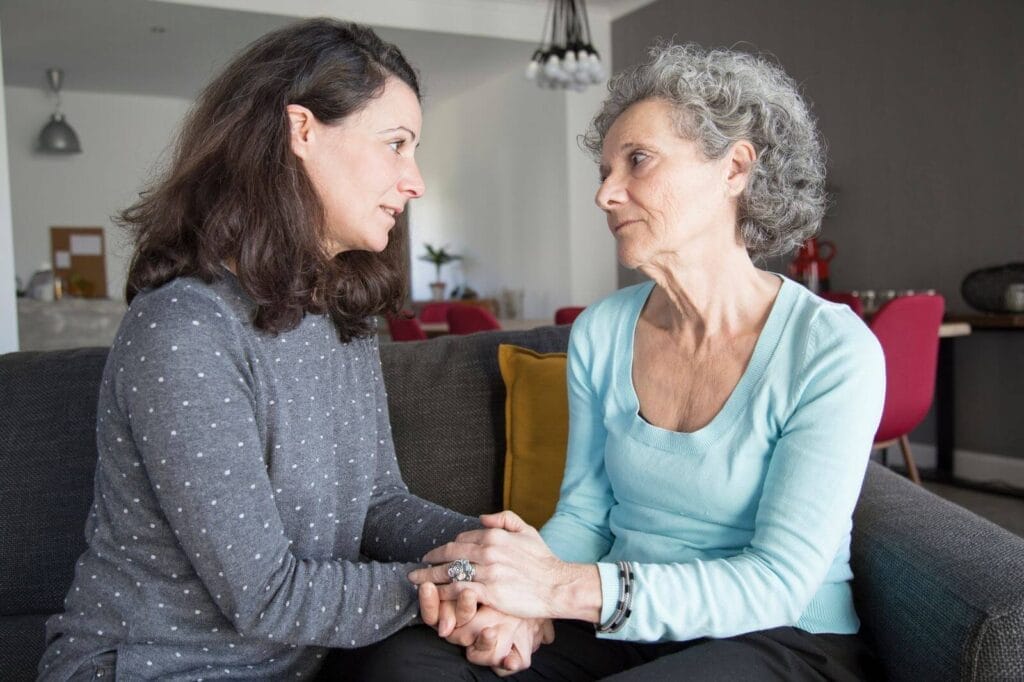 A woman sits on a couch with her older adult mother. They are holding hands.