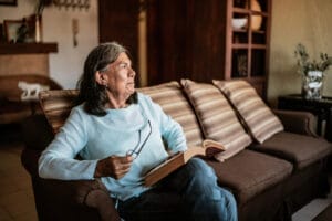 An older adult women sits on a couch. She looks up from the book she is reading, holding glasses in her hand.