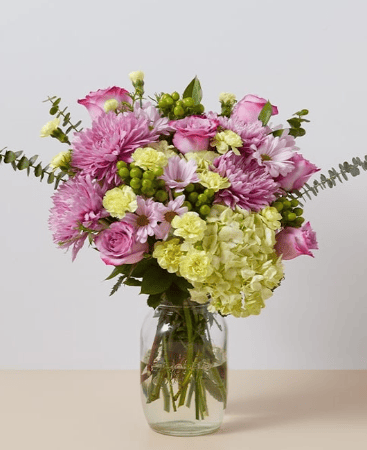 A bouquet of light purple and off-white flowers with green accent foliage in a jar-like vase sits on a tan table in front of a white backdrop.
