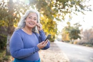 A woman is outside walking. She is putting an earbud in her ear and holding a smartphone.
