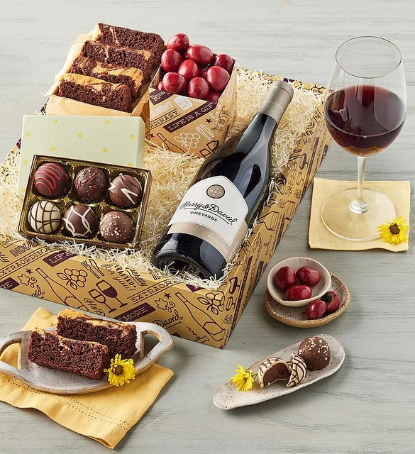 Product image of a gift box that holds slices of marble cake, chocolate truffles, chocolate-covered cherries, and a bottle of red wine. There is a glass of wine and plates with the food items next to the box.