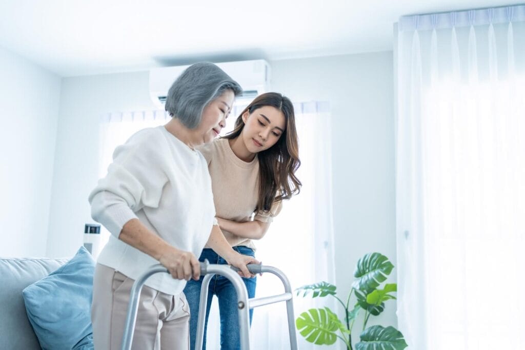 A woman helps her older adult mother walk using a walker.