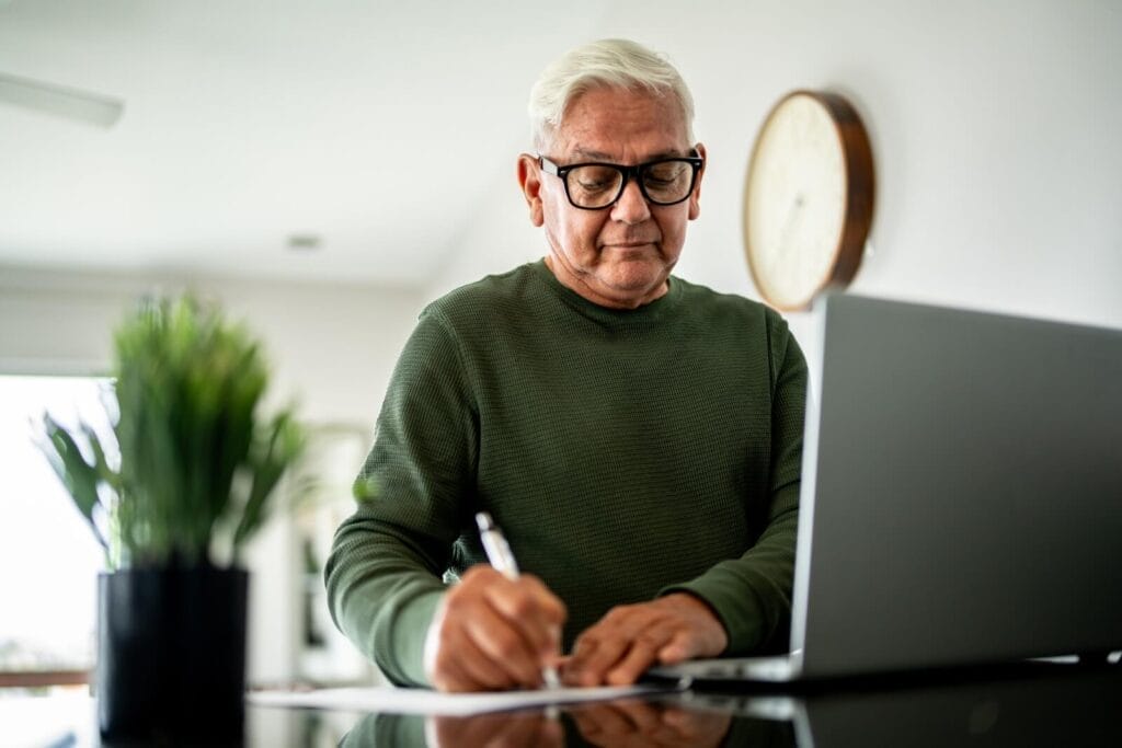 An older adult man is writing on a piece of paper. There is a laptop in front of him.