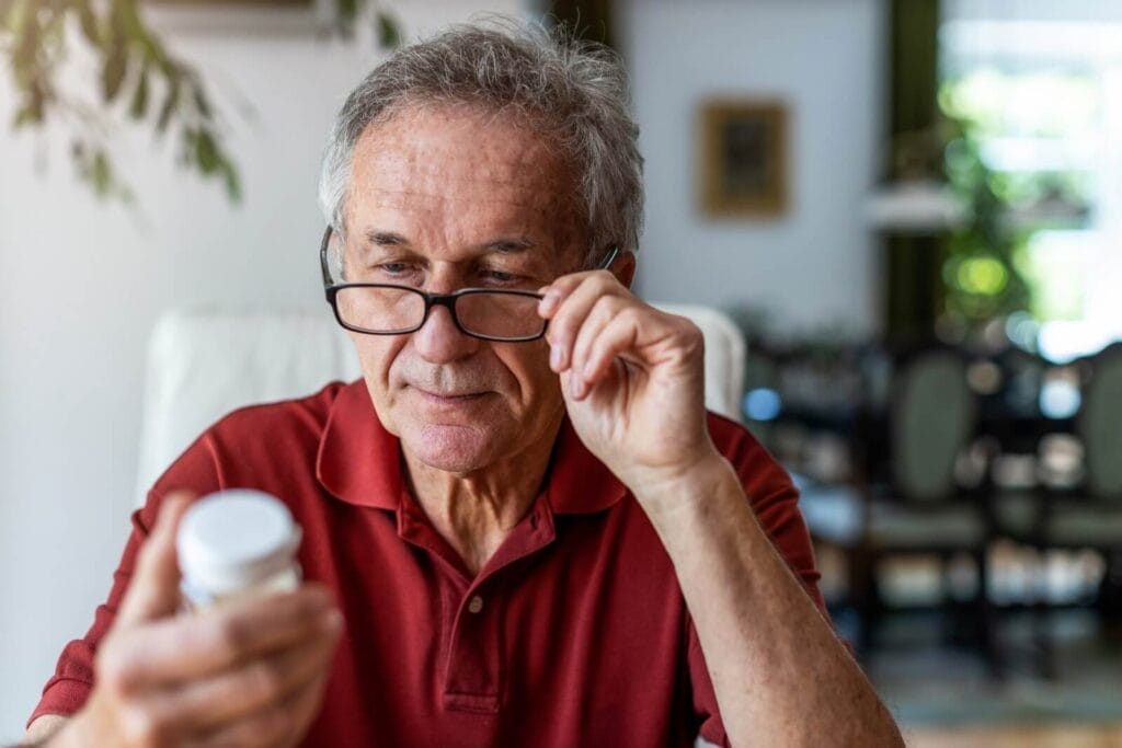 An older adult man adjusts his reading glasses as he examines a bottle of medication.